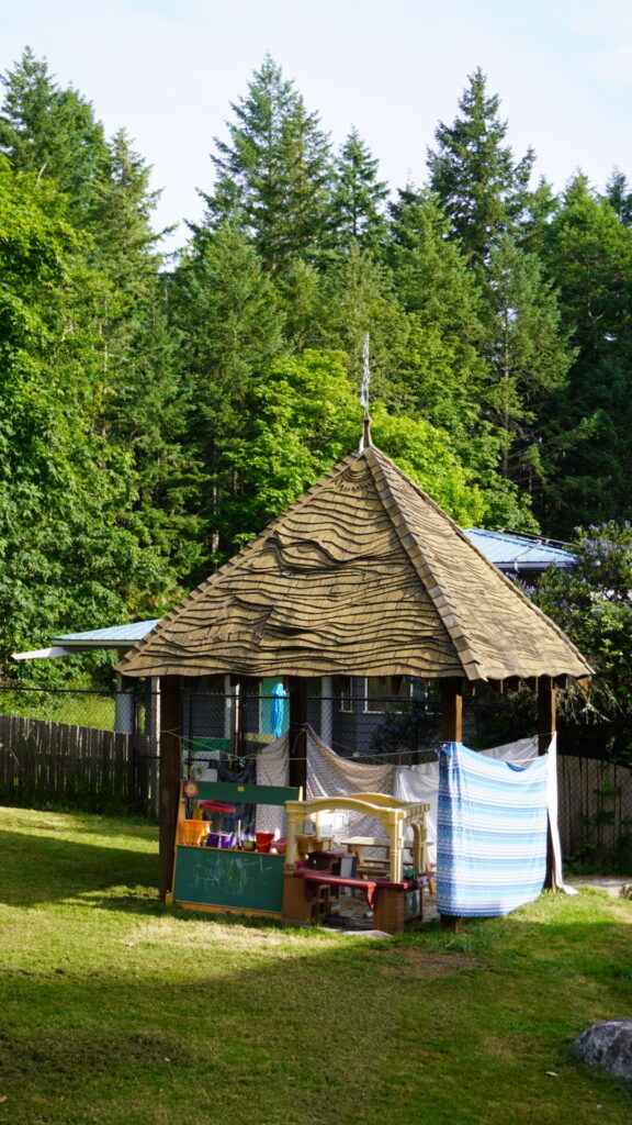 Wooden gazebo in a grassy play area, furnished with toy kitchen equipment and chalkboard, surrounded by trees and makeshift fabric walls.